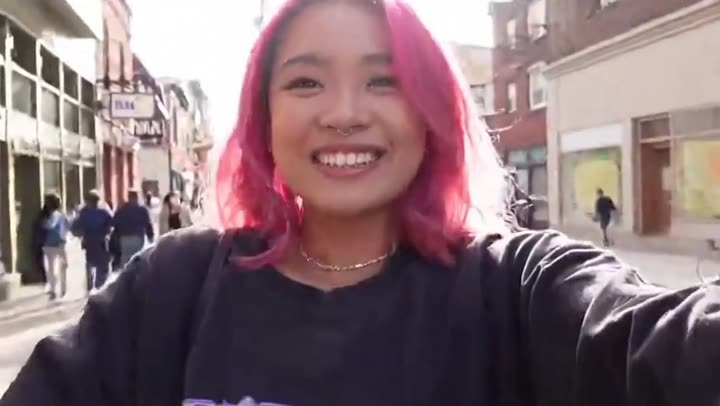A young woman with vibrant pink hair, wearing an oversized graphic t-shirt and ripped jeans, holds her phone out for a selfie vlogging her day of city exploration. She smiles warmly at the camera, her eyes sparkling with excitement. The background shows a bustling city street with diverse architecture and lively pedestrians. cut[0-4s]: She turns the camera to capture a quick pan of a colorful street art mural. cut[4-6s]: She bites into a steaming bao bun, her expression a mix of delight and slight messiness, with a busy market stall behind her. cut[6-9s]: The camera briefly focuses on her interaction with a friendly street vendor, both laughing. cut[9-12s]: She looks directly into the camera, a genuine smile on her face, sharing her experience, 'This city is amazing, I could explore forever!' Ambient city sounds, cheerful background music, and her enthusiastic narration fill the audio. Natural, bright daylight lighting. Casual, candid style.