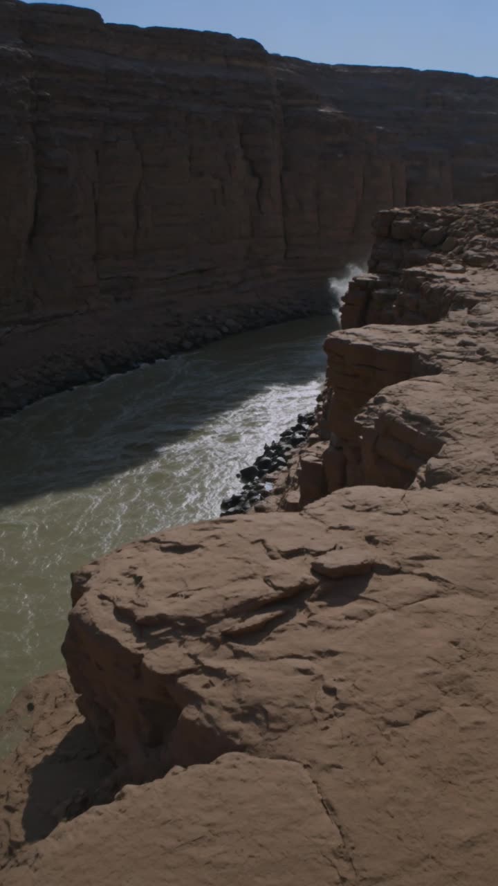 POV shot from a rocky shoreline next to a deep, fast-flowing river at the bottom of a desert canyon at dusk. Handheld camera with extreme wobbling. The operator zooms in rapidly on a massive displacement of water upstream. The enormous, multi-legged, armored back of a giant centipede-like anomaly breaches the surface, sliding down the canyon wall and displacing tons of water that crash against the rocks. The camera shakes as if the operator is stumbling backward in fear, the zoom losing and regaining focus on the moving armor plates. High-contrast shadows, amateur documentary style, photorealistic, massive scale.